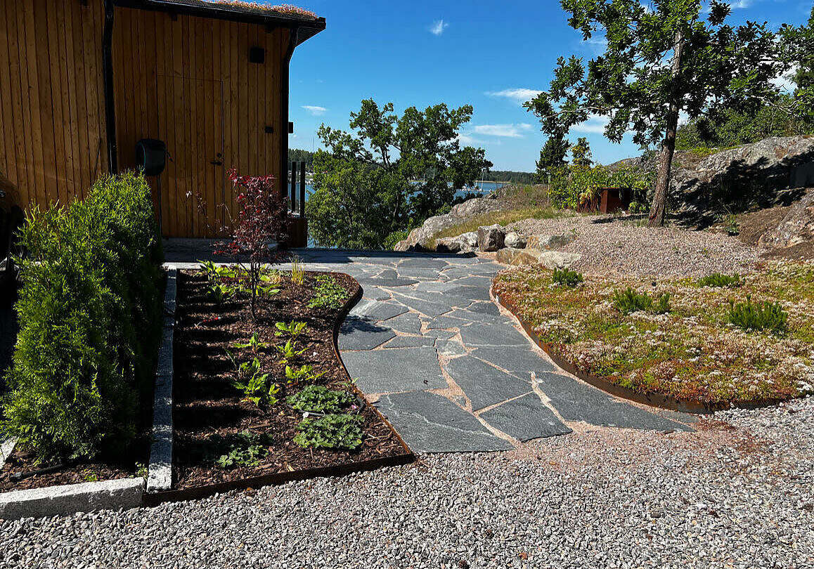 Foto på plantering med japansk lönn samt oregelbunden skiffer, kantstål av cortén och en plantering med sedummatta. Design av Östgöta trädgårdsdesign.